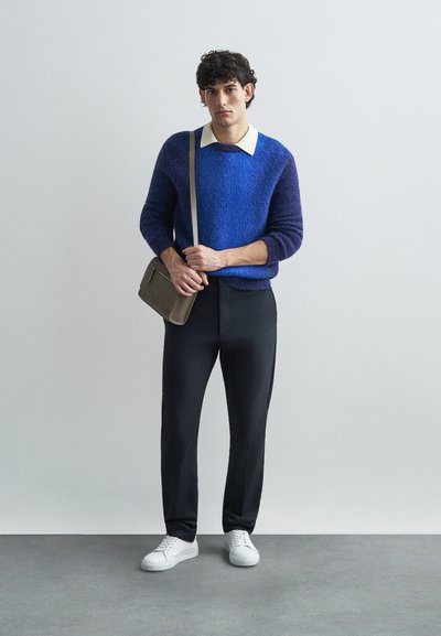 Suéter roxo de malha com colarinho creme, combinado com calças pretas e ténis brancos. A segurar uma mala cinza com detalhe em fecho.