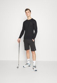 Black long-sleeve shirt, gray shorts with logo, holding a golf club, wearing white athletic shoes with blue accents and white socks.