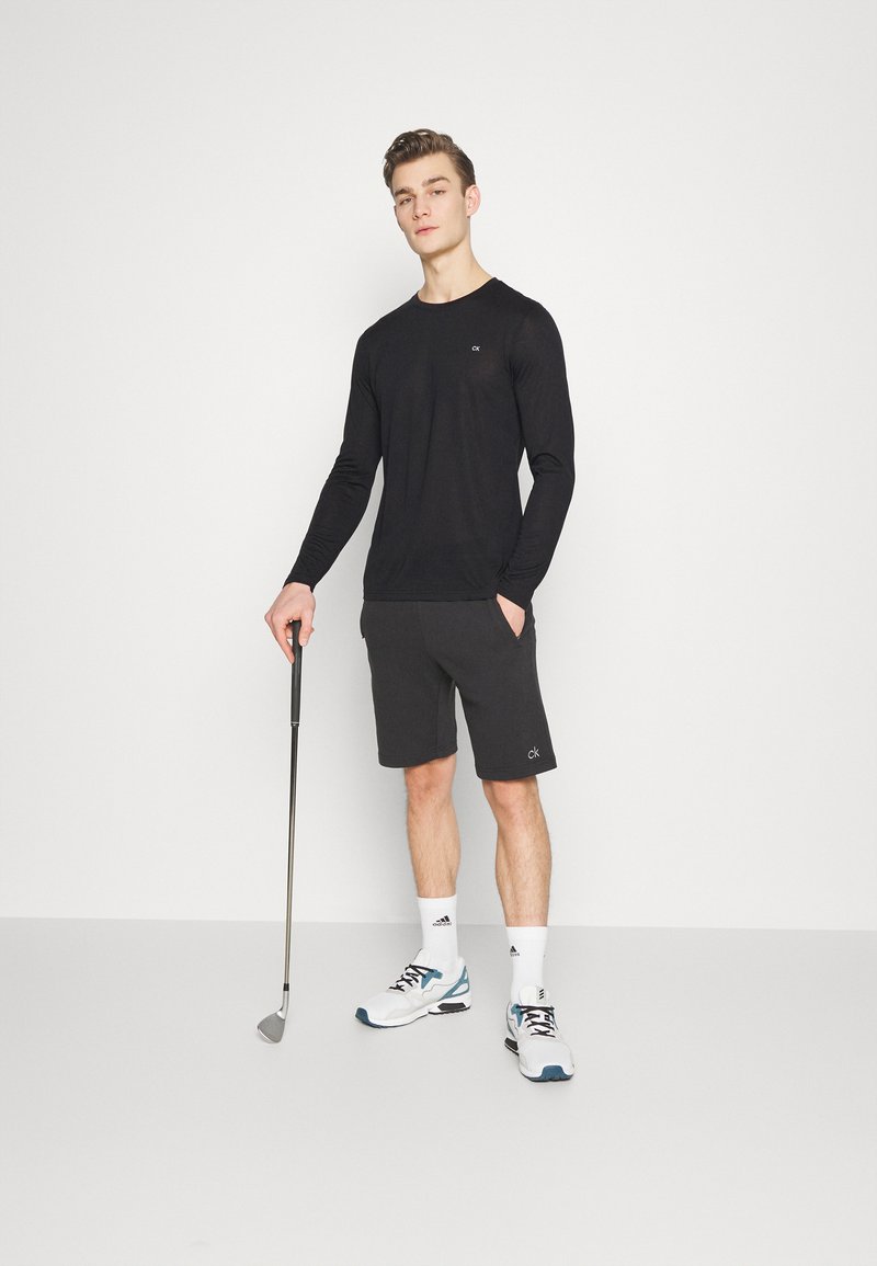 Black long-sleeve shirt, gray shorts with logo, holding a golf club, wearing white athletic shoes with blue accents and white socks.