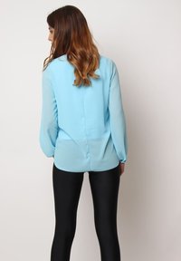 Blusa de manga larga de color azul claro con una textura suave y una costura en la parte posterior. Combinada con leggins negros brillantes. Fondo liso.