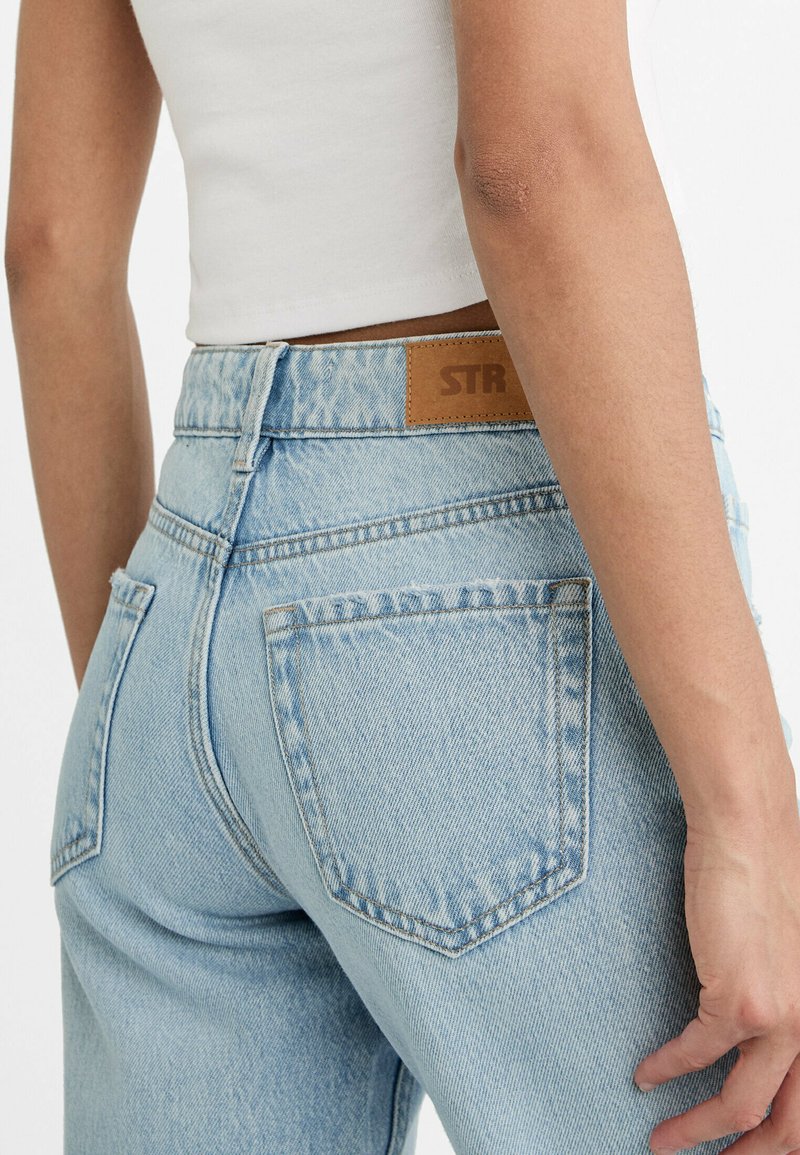 Jeans in denim azzurro chiaro con vita alta, dotati di una toppa in pelle marrone con l'etichetta "STR" e due tasche posteriori con dettagli cuciti.