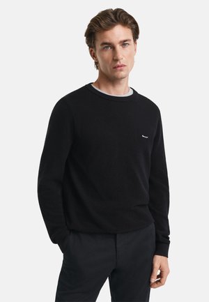 Zwarte textuur gebreide trui met een ronde hals, voorzien van een grijze binnenkraag en een klein logo op de borst. Gecombineerd met donkere broek.
