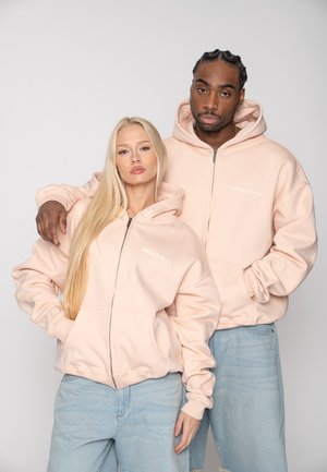 Twee jongvolwassenen dragen lichtroze hoodies met rits en lichtblauwe jeans, poserend tegen een effen grijze achtergrond.