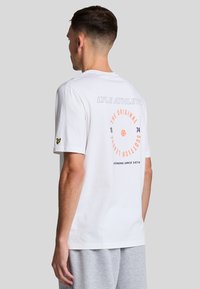 Camiseta de algodón blanca con un gráfico circular, texto en rojo y gris, y un acento del logotipo amarillo en la manga izquierda. Corte estándar.