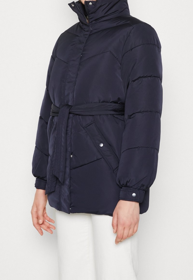 Navy puffer bunda s vysokým límcem, páskem v pase a kapsami na cvoky. Má prošívanou texturu a objemové rukávy.