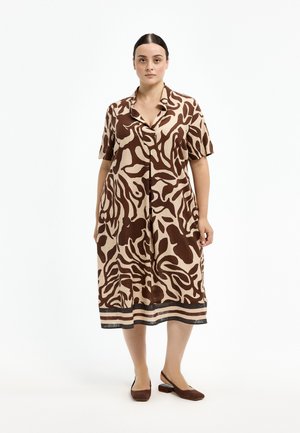 Frau steht und trägt ein knielanges, kurzärmeliges Kleid mit einem braun-beigen abstrakten Muster und passenden braunen flachen Schuhen.