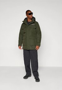 Žalia vandeniui atspari striukė su keliais kišenėmis, užtrauktuku ir gobtuvu. Dėvima su juodomis kargo kelnėmis ir tvirtais juodais batais.