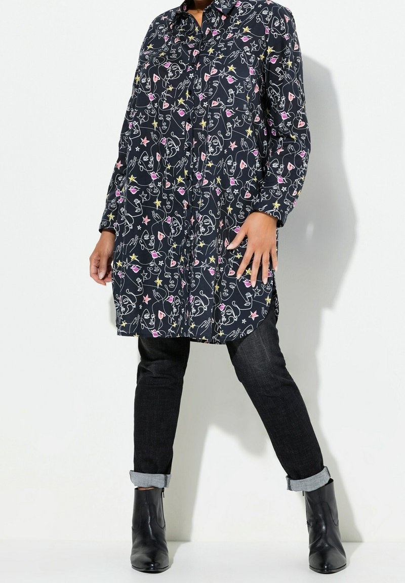 Personne portant une chemise noire à manches longues avec des motifs abstraits colorés de visages et d'étoiles, un jean noir avec revers et des bottines noires.