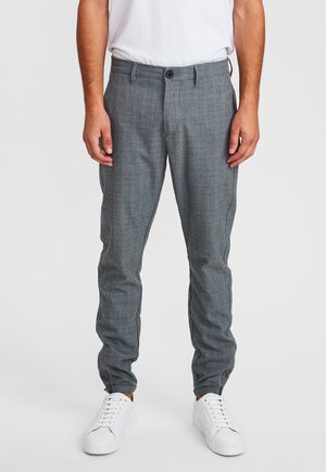 Mann trägt slim-fit graue karierten Hose und weiße Sneaker, steht vor einem schlichten hellen Hintergrund.
