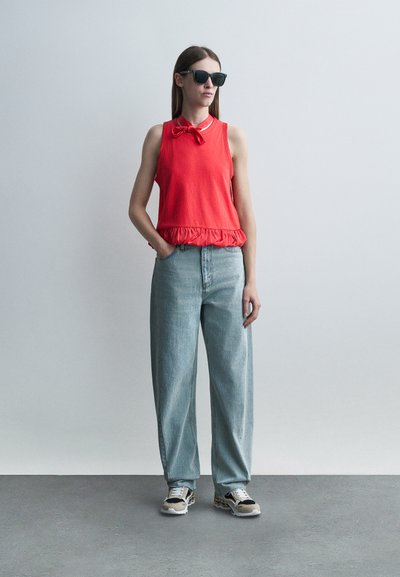 Γυναίκα στέκεται μπροστά σε απλό φωτεινό τοίχο, φοράει μαύρα γυαλιά ηλίου, αμάνικη κόκκινη μπλούζα με φιόγκο, άνετο ανοιχτό μπλε τζιν και αθλητικά παπούτσια.