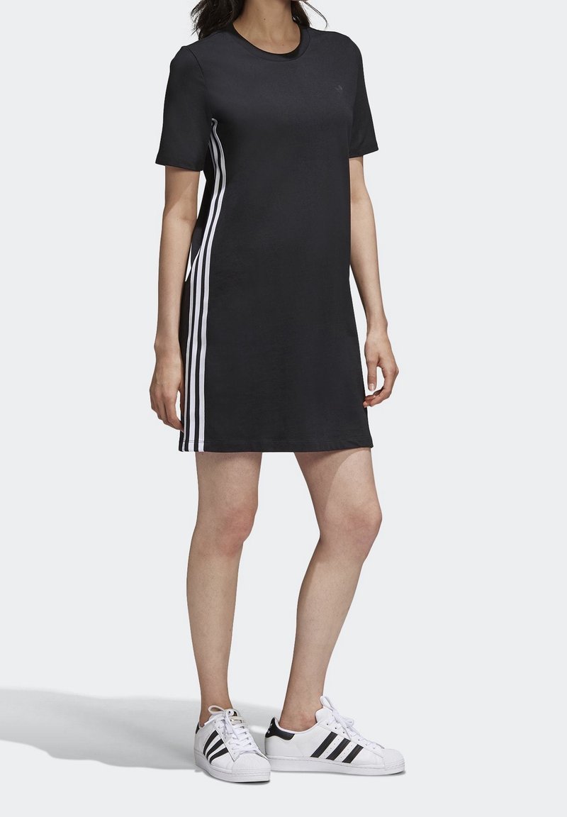 Kvinna iklädd en svart kortärmad klänning med vita sidostråk och vita sneakers med svarta stråk, ståendes mot en vit bakgrund.