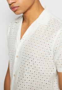 White short-sleeve shirt made of textured fabric with circular eyelet patterns and a relaxed collar, featuring a button closure.