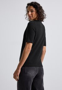 Kvinde med krøllet hår iført en sort ribbet kortærmet top og bløde sorte jeans, der ser over sin skulder foran en ensfarvet baggrund.