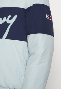 Pružná bunda ve světle modré a námořnické barvě, s velkým bílým logem přes hruď, bočními kapsami a emblémem Tommy Hilfiger na rukávu.