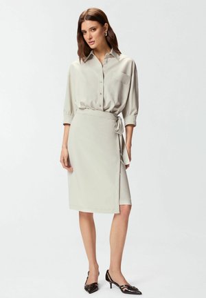 Donna con camicia color beige chiaro con bottoni e maniche a tre quarti e gonna a portafoglio abbinata, che indossa décolleté nere con punta a punta, in piedi davanti a uno sfondo bianco.