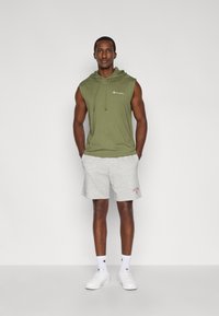 Αμάνικο φούτερ με κουκούλα στο χρώμα ελιάς με κορδόνια, συνδυασμένο με ανοιχτό γκρι σορτς. Λευκά αθλητικά παπούτσια και κάλτσες με λεπτομέρεια λογότυπου.