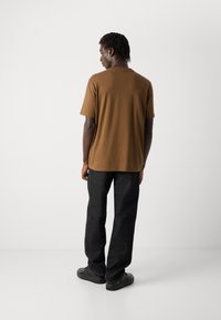 Ein Mann steht mit dem Rücken zur Kamera, trägt ein braunes Kurzarmhemd, schwarze Hosen und schwarze Turnschuhe vor einem schlichten weißen Hintergrund.