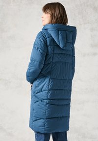 Blå vadderad lång jacka med huva, har en quiltad design, slät yta och sidofickor, bärs över mörka jeans.