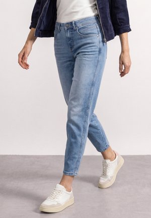 Personne portant un jean bleu clair, des baskets blanches, une veste bleu marine et un haut blanc marchant sur un sol gris avec un fond blanc uni.