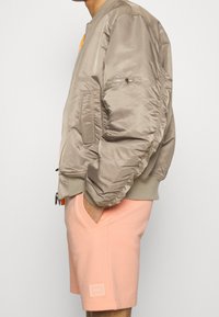 Veste bomber en satin beige clair avec des poignets côtelés, poche zippée sur le bras et accent orange sur la poitrine, assortie à un short rose.