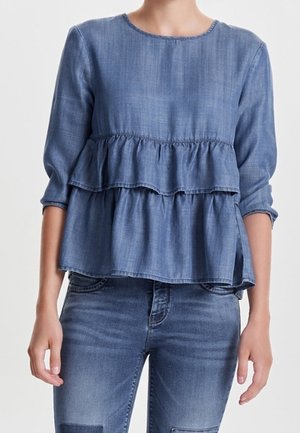 Kobieta ubrana w długi rękaw peplum z niebieskiego denimu w połączeniu z niebieskimi dżinsami, stojąca na tle jasnego, gładkiego tła.