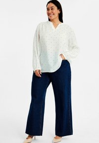 Vit långärmad blus med svarta prickar, v-ringning och något rynkade manschetter, tillsammans med vida mörkblå jeans och beiga skor.