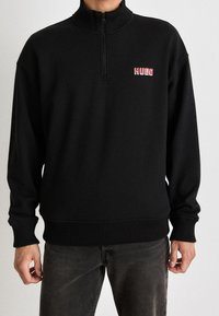 Uomo che indossa un maglione nero con zip a quattro quarti e polsini e orlo a costine, con il logo rosso "HUGO" sul petto, abbinato a jeans scuri.