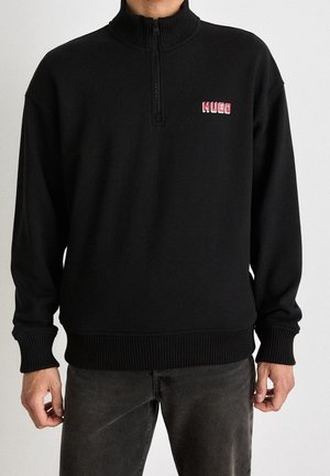 Hombre vistiendo un suéter negro con cremallera hasta el cuarto, con puños y dobladillo de canalé, y el logo rojo de "HUGO" en el pecho, combinado con jeans oscuros.