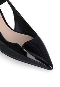 Puntale in vernice nera con punta a lancia e design con ritagli, dalla finitura liscia. Presenta una soletta beige e una silhouette elegante.