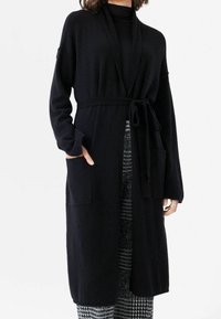 Lång svart cardigan i mjukt tyg, med bälte, öppen front och två framfickor. Ärmarna är vida och något texturerade.