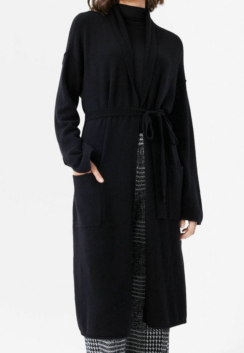 Lång svart cardigan i mjukt tyg, med bälte, öppen front och två framfickor. Ärmarna är vida och något texturerade.
