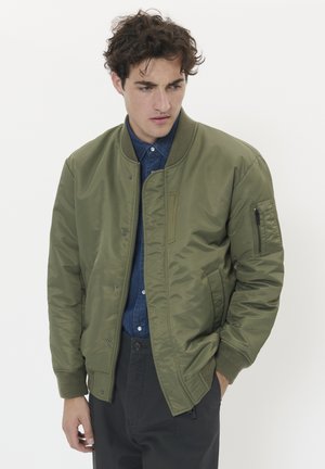 Olivenfarbene Bomberjacke mit Reißverschluss, Ribkragen und -bündchen, seitlichen Taschen und einer mit Reißverschluss versehenen Ärmeltasche. Über einem blauen Hemd getragen.