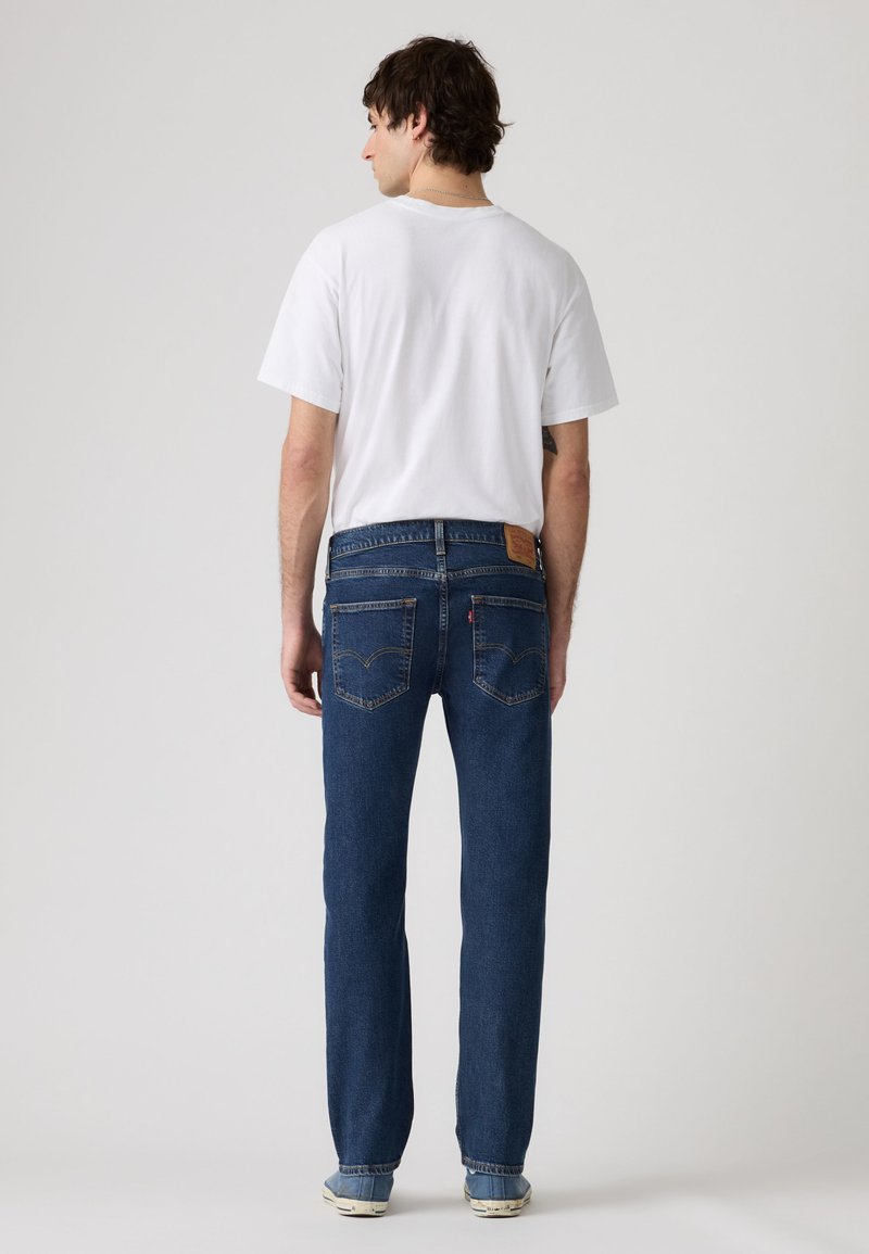 Uomo in piedi di spalle che indossa una maglietta bianca, jeans blu e scarpe da ginnastica azzurro chiaro su uno sfondo neutro.