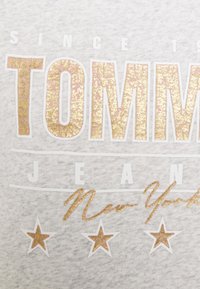 Camiseta de algodón gris con un texto audaz y brillante en dorado y rosa que dice "TOMMY JEANS" acompañado de estrellas decorativas y líneas.