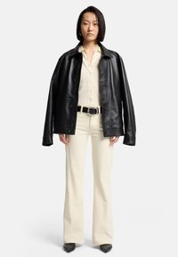 Sort oversized læderjakke over en cremefarvet skjorte med knapper, kombineret med cremefarvede flared bukser og spidse sorte sko; glat tekstur hele vejen igennem.