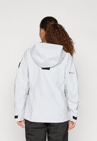 GORE-TEX-takki vaaleanharmaat väri, jossa on huppu, mustat yksityiskohdat, vetoketjulliset sivutaskut ja hieman pidempi selkäosa lisäsuojaa varten.