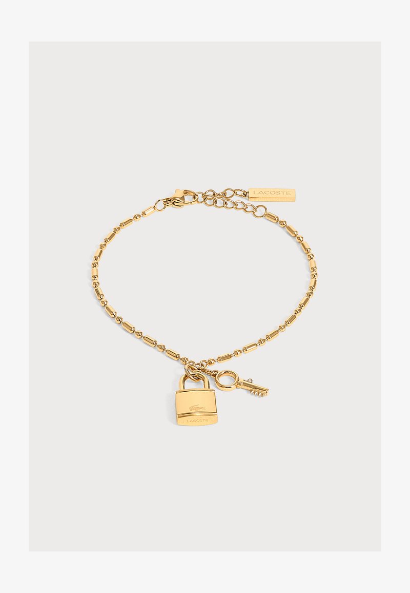 Bracelet chaîne à perles dorées avec breloques cadenas et clé, logo Lacoste sur le cadenas et étiquette rectangulaire près du fermoir.