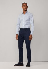 Biała koszula z niebieskimi wzorami, zapinana na guziki, zestawiona z granatowymi spodniami. Model nosi ciemne buty i pasujące skarpetki.