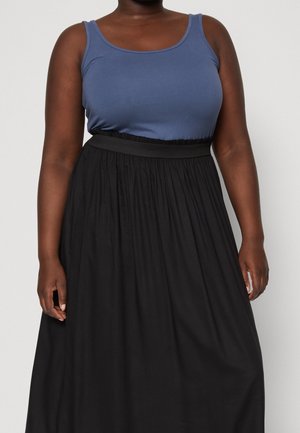 Femme portant un haut bleu sans manches et une jupe plissée noire taille haute, debout devant un fond clair uni.