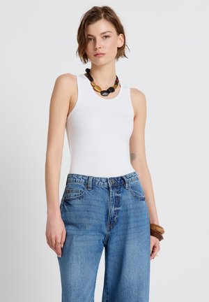 Giovane donna con top bianco senza maniche, jeans blu a vita alta, collana spessa nera e oro e braccialetti di legno, in piedi contro una parete bianca.