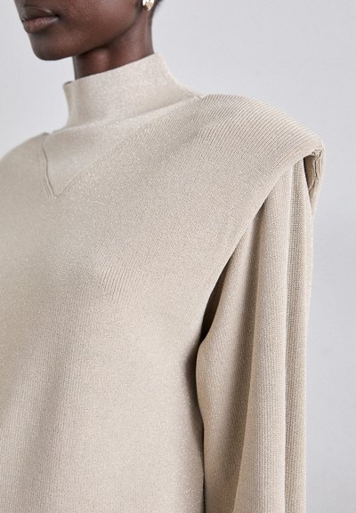 Beige højhalset sweater med ribbet tekstur, skulderpuder og subtil glans; har en lille V-udskæring og lange ærmer.