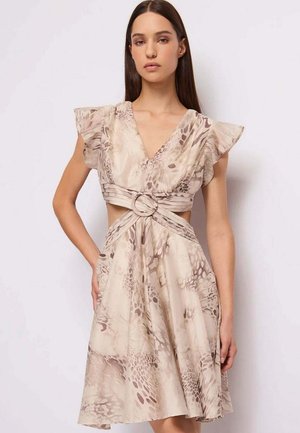 Donna che indossa un abito beige e taupe con stampa astratta, maniche a campana, scollo a V profondo e fibbia circolare nel taglio in vita, in piedi di fronte a una parete bianca.