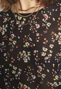 Svart blommig blus med små vita, gula och röda blommor, med en mjuk textur; lager av guldhalsband pryder halslinjen.