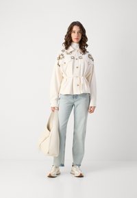 Giacca bianca ricamata con vita regolabile, abbinata a jeans azzurri chiari e sneakers beige, mentre tiene una borsa color crema.