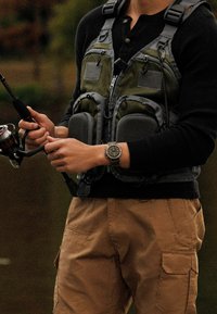 Chaleco de pesca en verde y gris con múltiples bolsillos; camiseta de manga larga negra; pantalones cargo beige; sosteniendo una caña de pescar con carrete.
