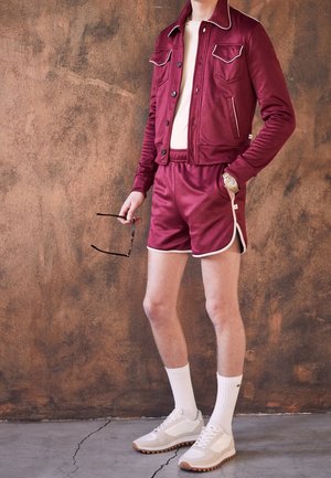 Persona con chaqueta burdeos y pantalones cortos a juego con ribete blanco, calcetines blancos, zapatillas beige, sosteniendo gafas de sol frente a una pared marrón con textura.