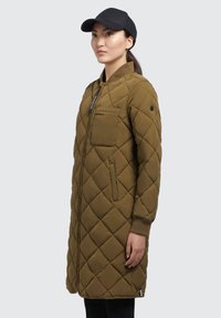 Manteau long matelassé vert olive avec fermeture éclair, poignets côtelés et poche poitrine. Porté avec une casquette noire sur un fond neutre.