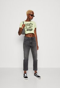 Przycięta limonkowa koszulka z czarnym nadrukiem, szare dżinsy o wysokim stanie i czarne trampki z białymi sznurówkami; niesie szarą torebkę.