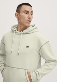 Sudadera verde claro hecha de tejido suave. Cuenta con un bolsillo canguro, cordones y costuras en contraste. Pequeño logo en el pecho.