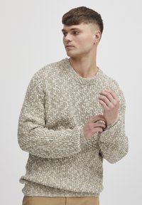 Beige knit sweater with a textured pattern, ribbed cuffs, and crew neckline, worn by a model with short hair in a neutral background.